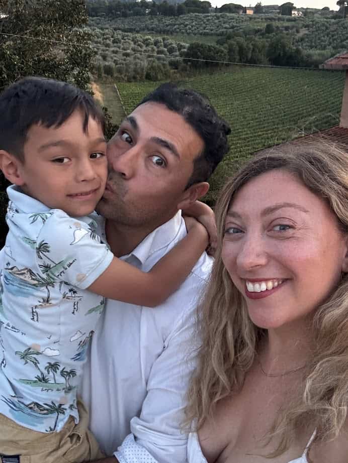 A smiling woman takes a selfie with a man and a child in front of green fields at sunset, capturing memories from their 4 days in Castel Gandolfo.