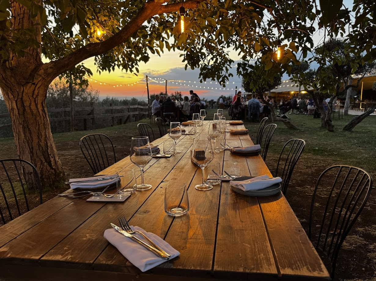 A long wooden table set for dinner under string lights at sunset, with people dining in the background&mdash;perfect for savoring evenings during 4 days in Castel Gandolfo.