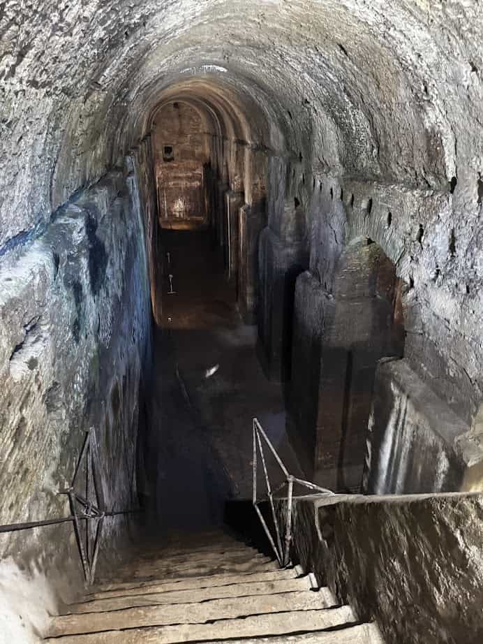 Stone stairs descend into an ancient, arched underground tunnel with pillars and dim lighting—a hidden gem to discover during 4 days in Castel Gandolfo.