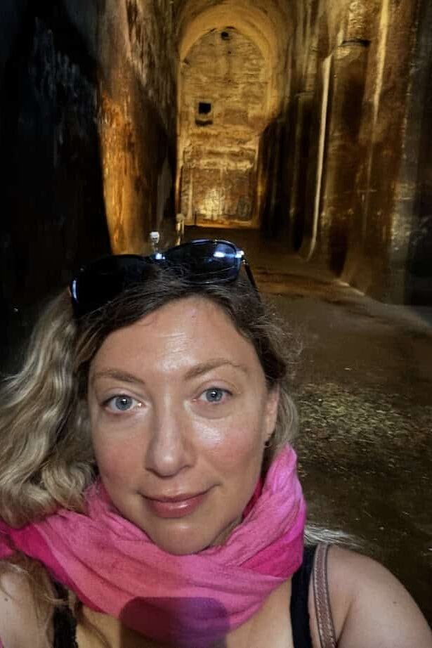 Woman with blonde hair and pink scarf takes a selfie in a dimly lit, ancient underground tunnel, capturing a moment from her 4 days in Castel Gandolfo.