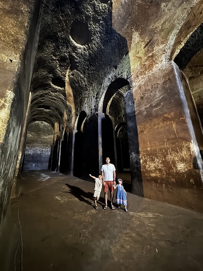 An adult and two children stand inside a large, ancient, arched stone chamber with high vaulted ceilings, exploring history as part of their 4 days in Castel Gandolfo.