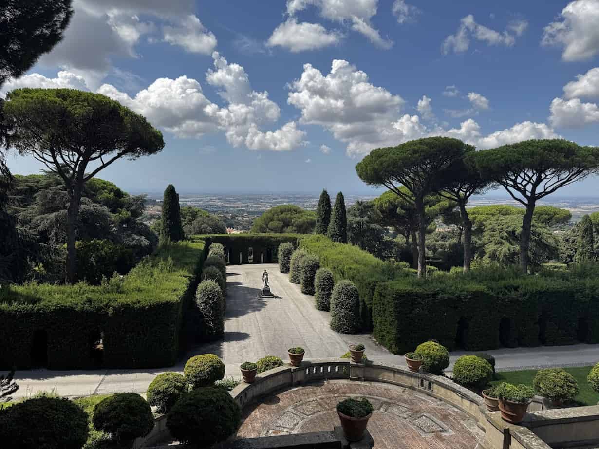 A sunny garden with tall trees, hedges, potted plants, and a statue under a blue sky with scattered clouds—perfect for relaxing after 4 days in Castel Gandolfo.