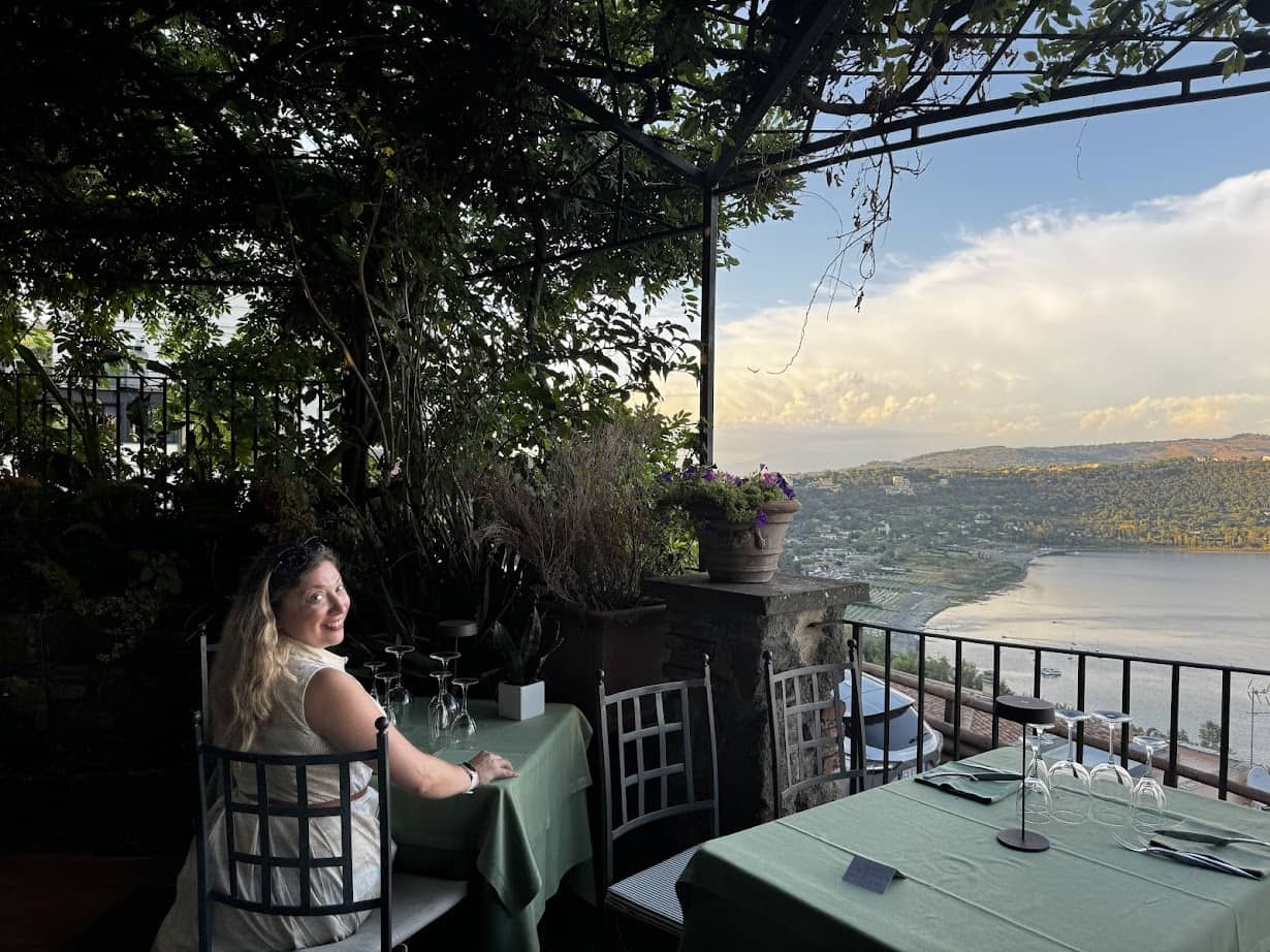 A woman sits at a table with a scenic view of a lake, hills, and sky from a terrace restaurant, savoring the beauty of 4 days in Castel Gandolfo.