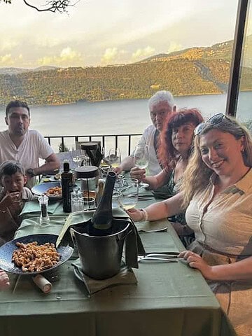 A family of six enjoys dinner at an outdoor lakeside restaurant with pasta and wine on the table, savoring their 4 days in Castel Gandolfo.