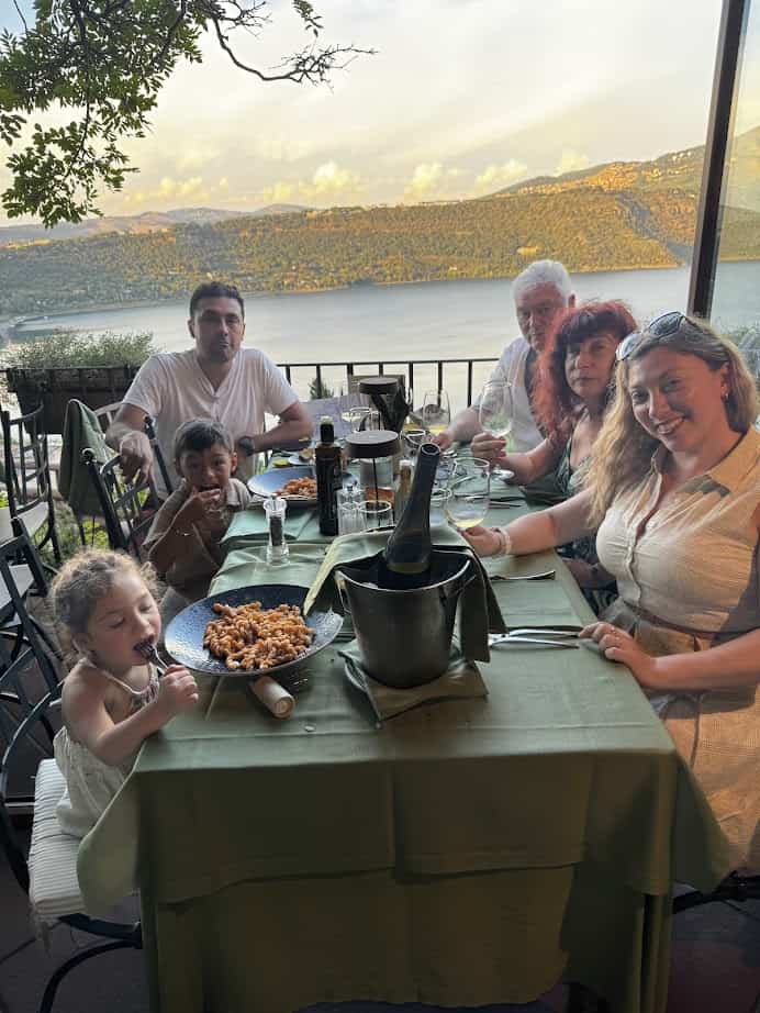 A family of six dines outdoors by a lake in Castel Gandolfo, enjoying food and drinks at a table with a scenic view during their 4 days in the charming town.