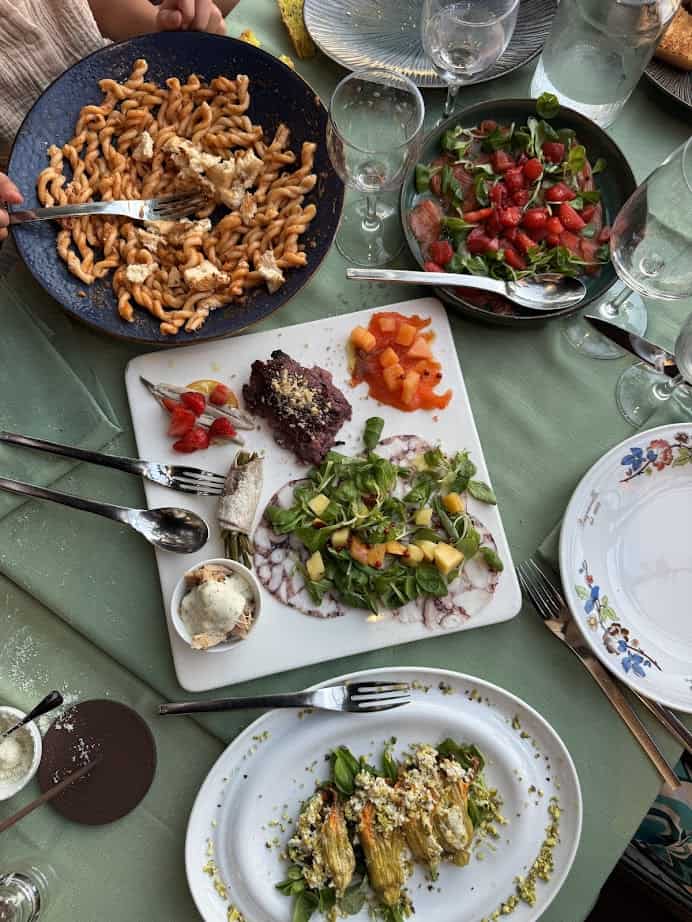 An overhead view of a table set for 4 days in Castel Gandolfo, featuring pasta, salads, appetizers, and empty wine glasses on a green tablecloth.