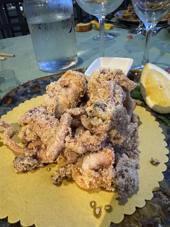 A plate of fried calamari on yellow paper, served with a lemon wedge and a glass of water in the background—perfect for enjoying during 4 days in Castel Gandolfo.