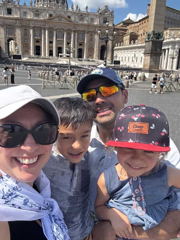 A smiling family of four poses in front of St. Peter’s Basilica on a sunny day, excited to explore the best gluten free restaurants in Rome.