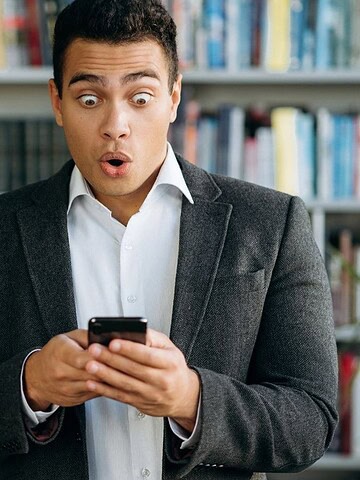 A surprised man in a suit looks at his phone while standing in front of bookshelves in a library.