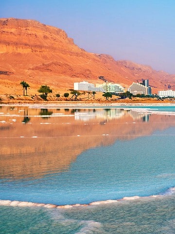 Turquoise water and salt formations along a coast with hotels and mountains in the background under a clear sky.