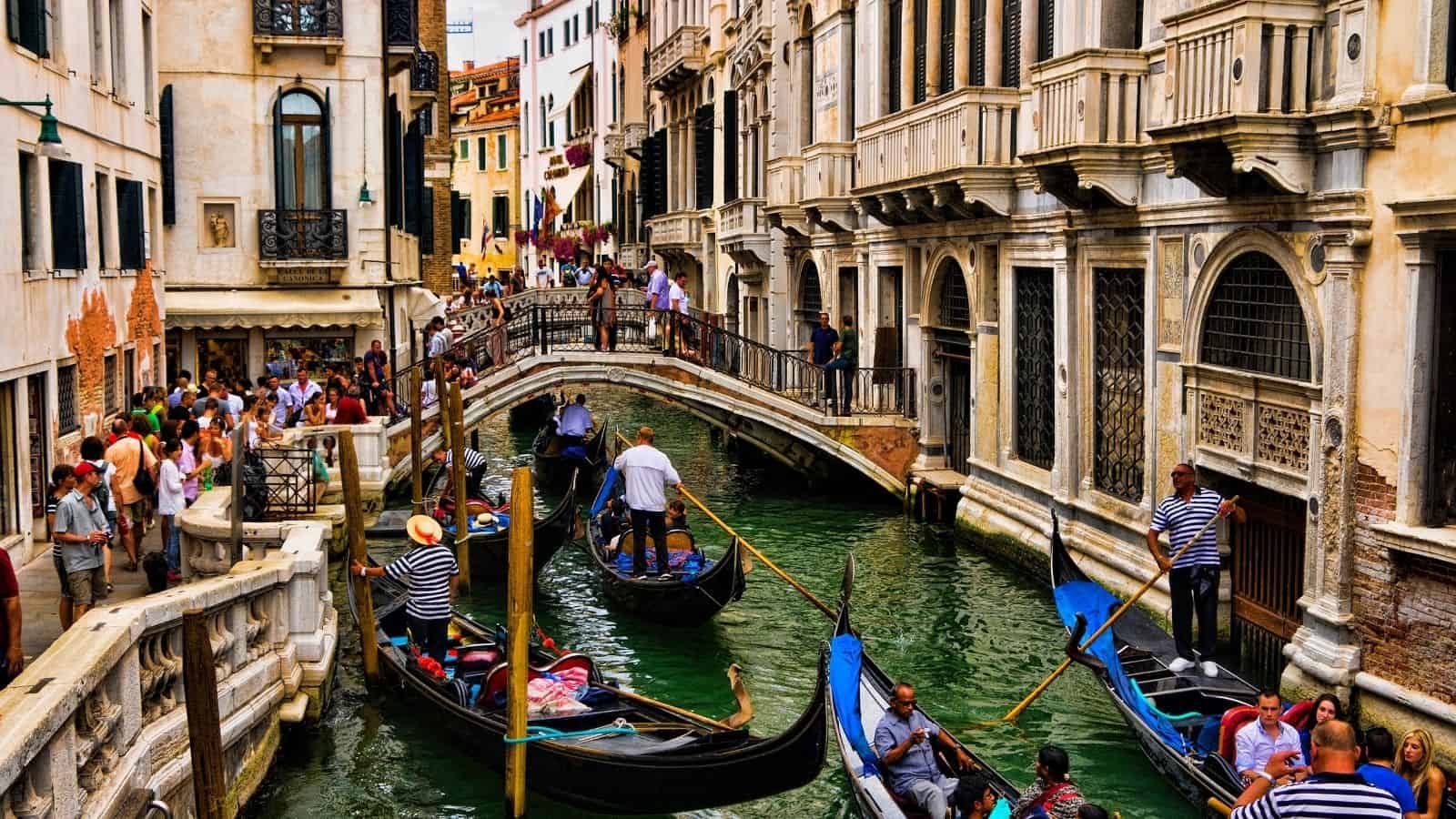 Crowded Venice canal with gondolas, people on a bridge, and historic buildings lining the water.