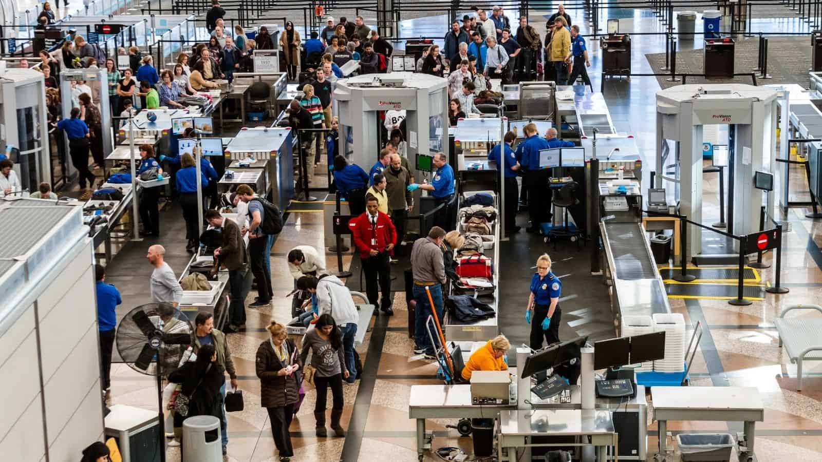 A busy airport security checkpoint with travelers in line and TSA agents screening bags and people.