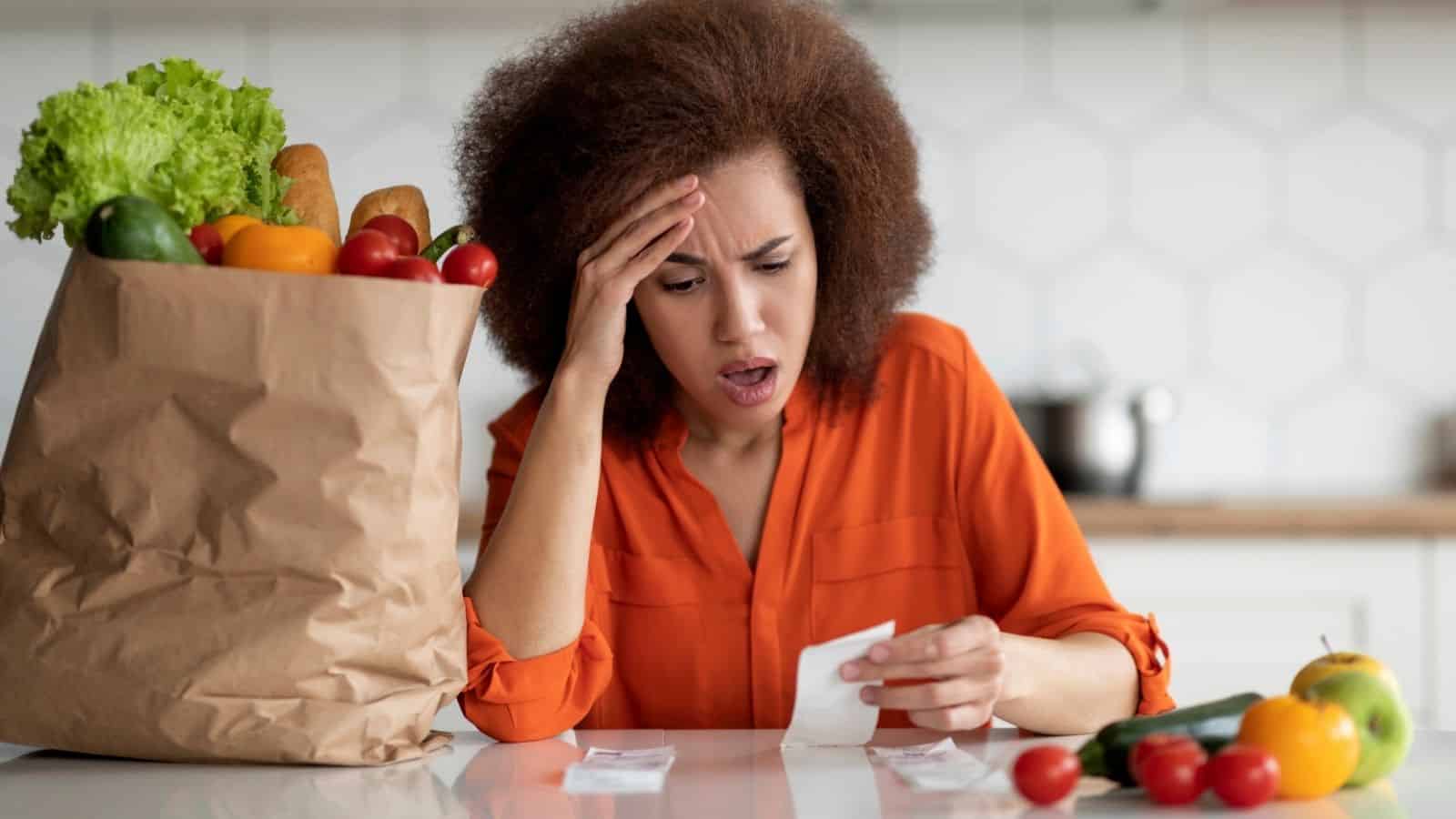 Woman looking stressed at a grocery receipt next to a bag of groceries on a kitchen counter.