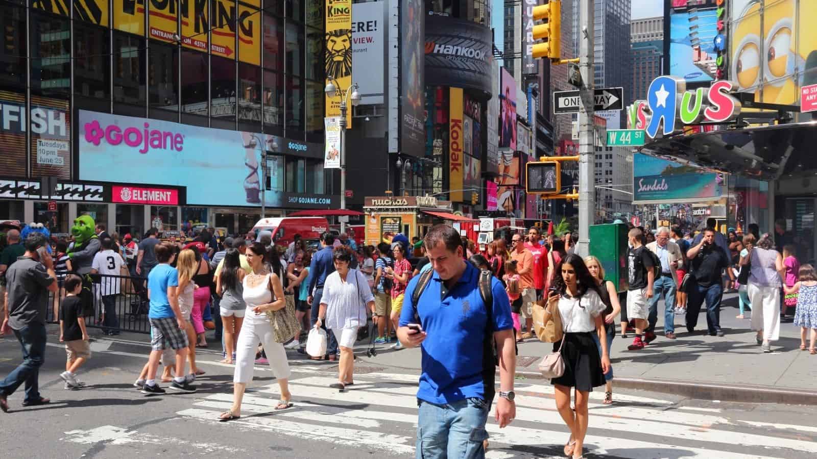 Crowded city street with people crossing at a crosswalk; bright billboards and shops in the background.