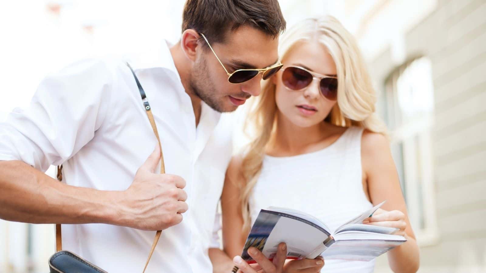 A man and woman wearing sunglasses look at a guidebook together while standing outdoors.