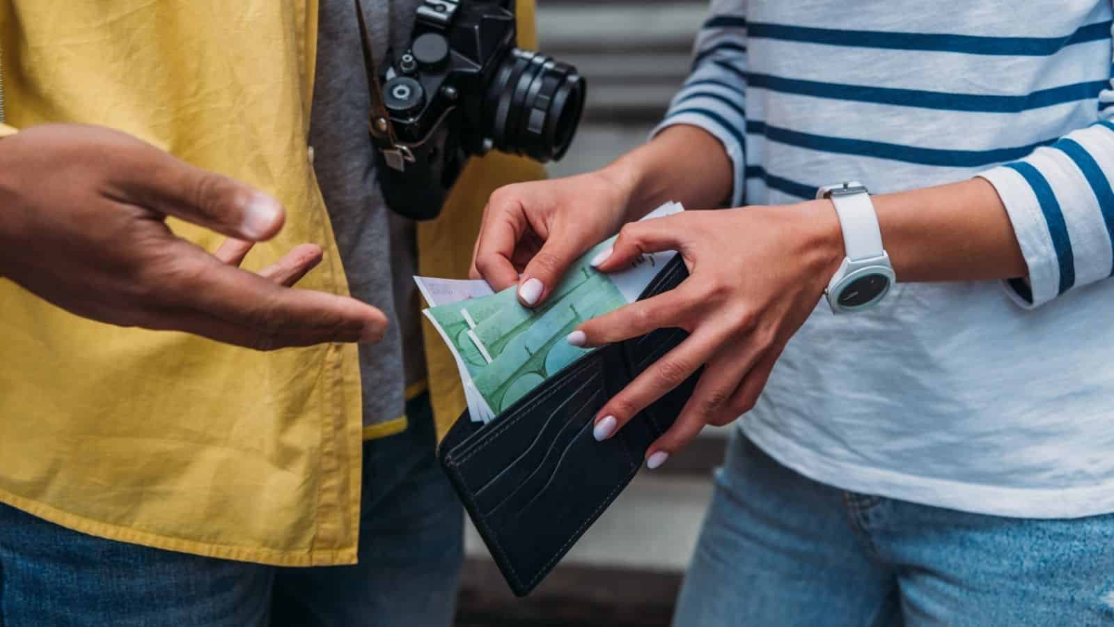 Two people exchange money&mdash;one holds a black wallet filled with cash and tickets, ideal for budget travel, while the other gestures with an open hand.