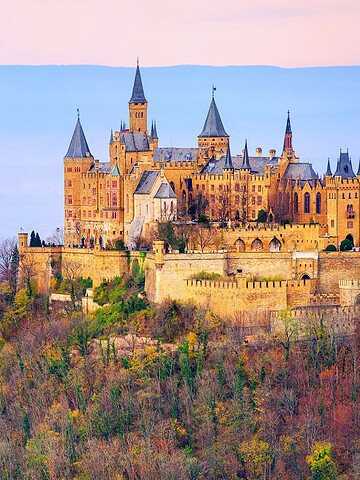 Hilltop castle with tall towers surrounded by colorful autumn trees and misty blue mountains in the background.