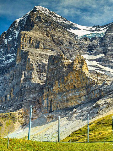 A red train travels along tracks at the base of a steep, rocky mountain with patches of snow and green grass.