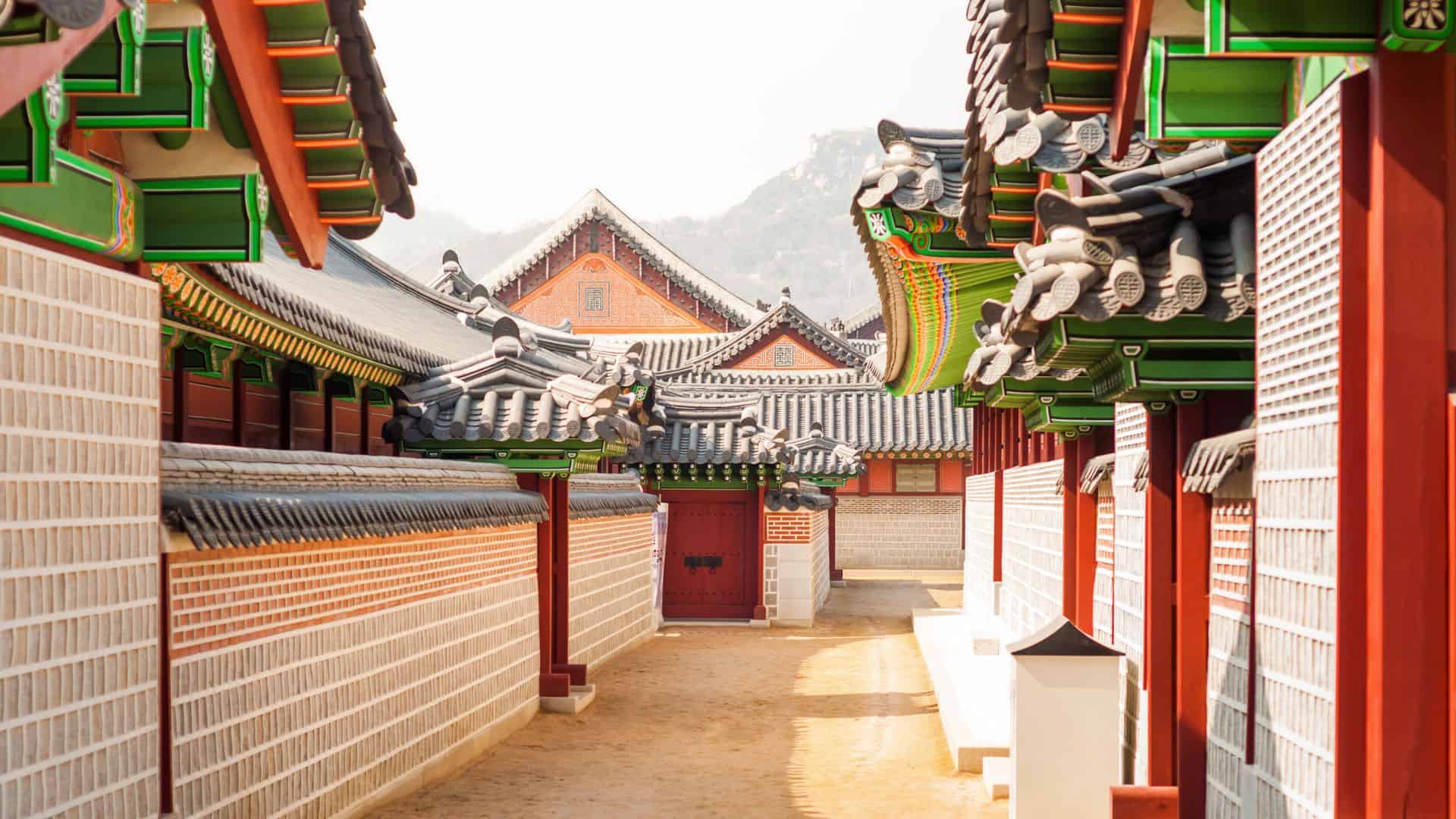 Traditional Korean palace buildings with ornate roofs and a sunlit walkway between them.