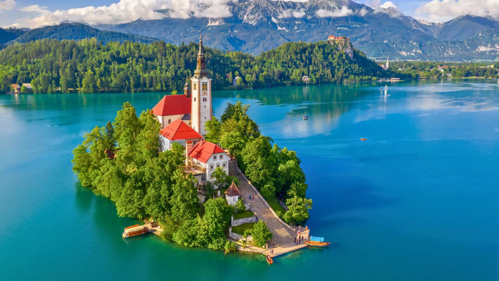 A small island with a church and red roofs on a blue lake, surrounded by green hills and distant mountains.