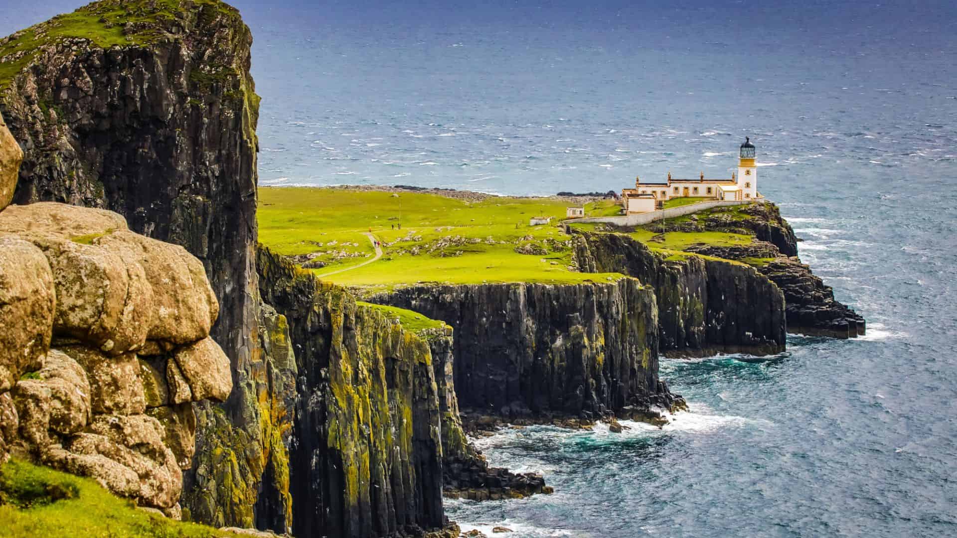 A lighthouse sits on dramatic green cliffs overlooking the ocean under a cloudy sky.