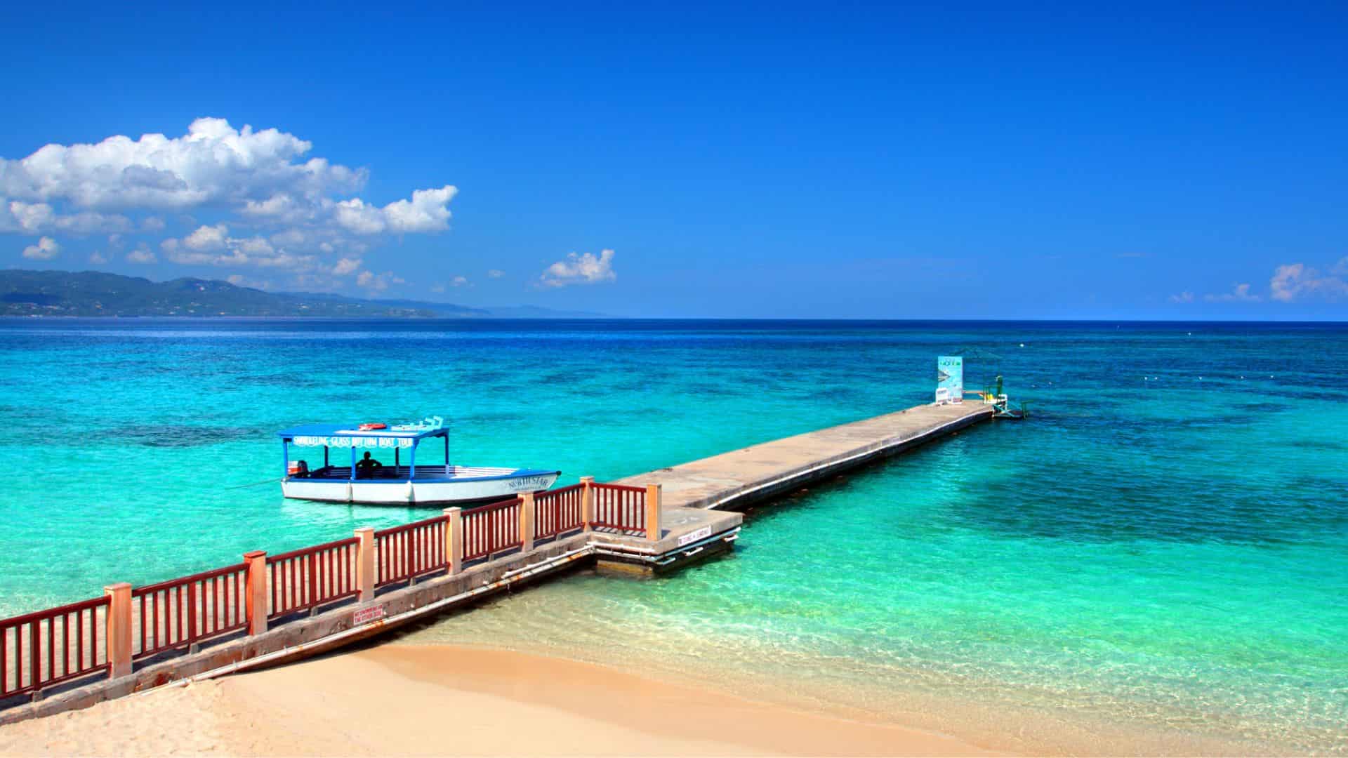 A wooden pier extends into clear turquoise water with a small boat docked and mountains in the distance.