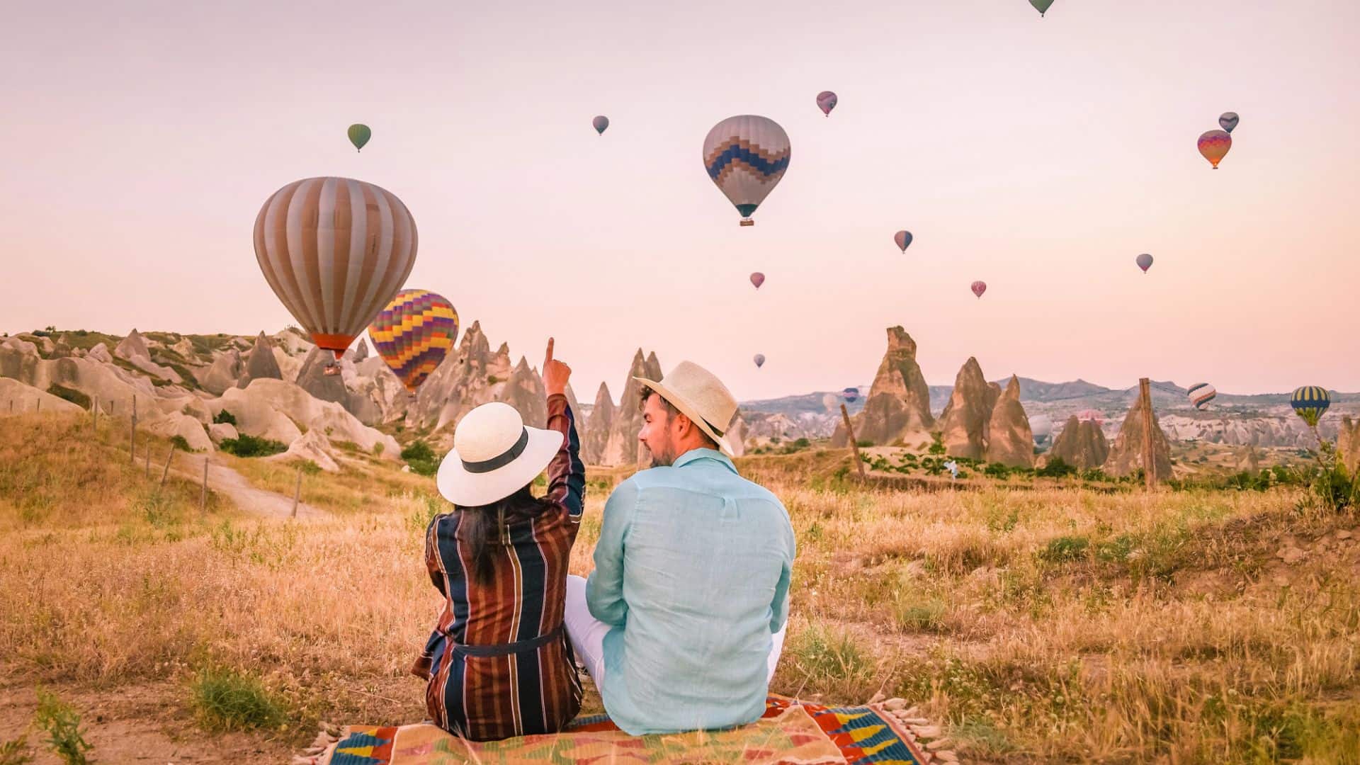A couple sits on a blanket in a field, watching hot air balloons float over rocky formations at sunrise.