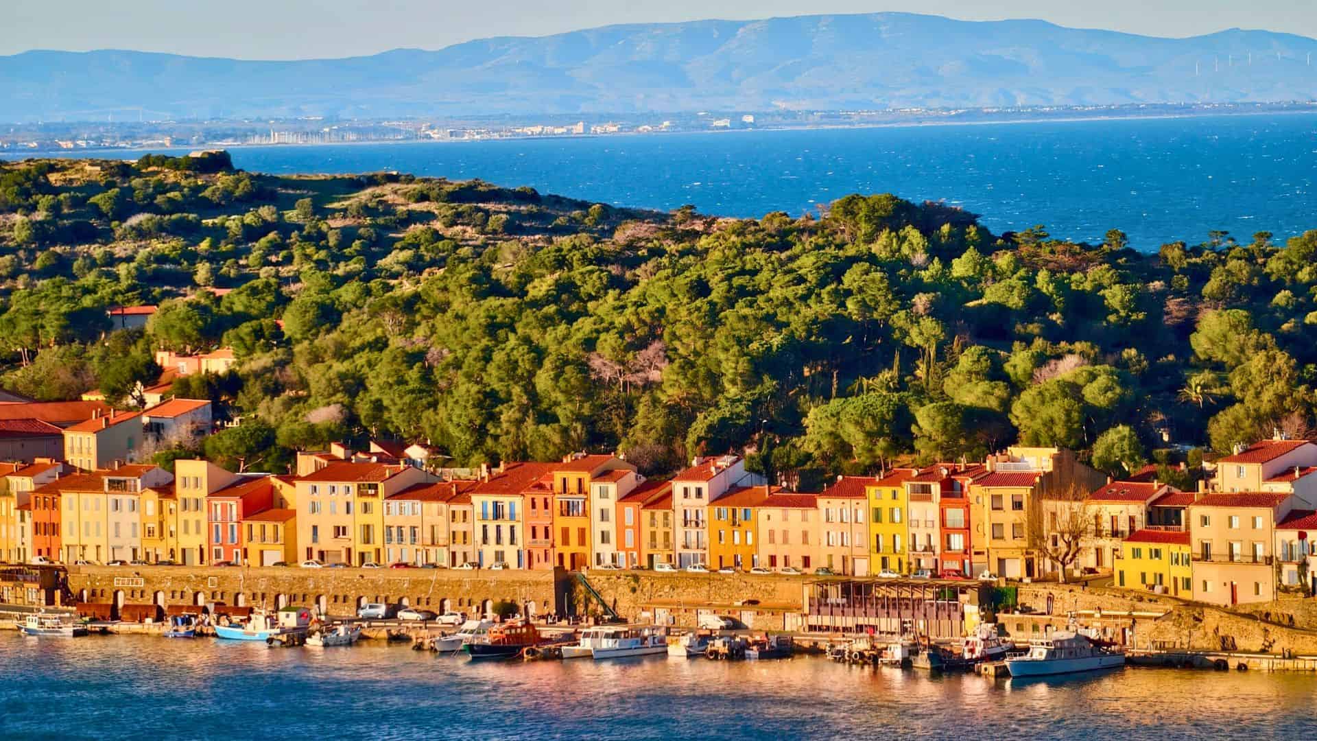 Colorful houses line a waterfront with boats docked, lush green hills, and mountains in the background.