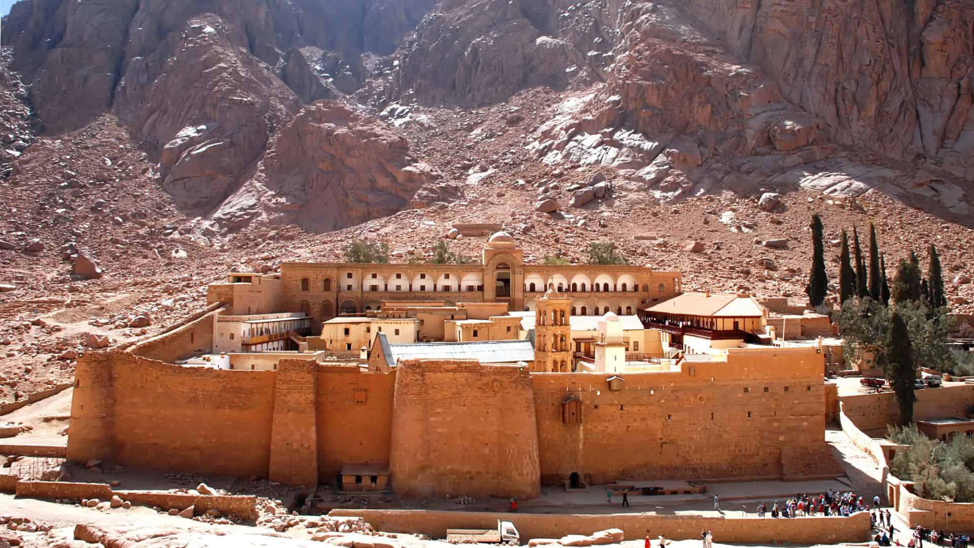 Ancient stone monastery with high walls, nestled at the base of rugged, rocky mountains in a desert landscape.