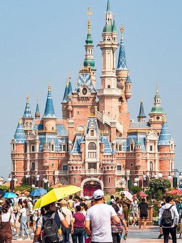 Crowds of people walk toward a large Disney castle on a sunny day, surrounded by green trees.