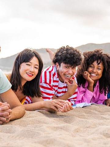 Six young adults lying on the sand at the beach, smiling and enjoying a sunny day together.
