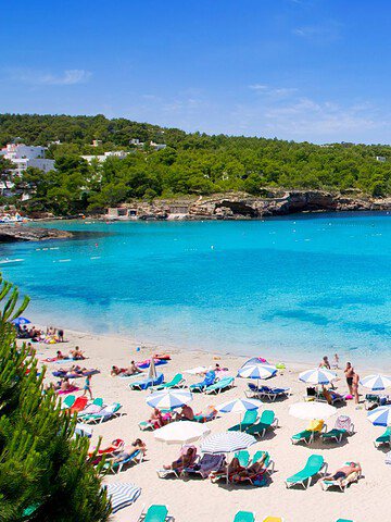 Crowded sandy beach with turquoise water, sunbathers on loungers, and boats anchored in the bay.