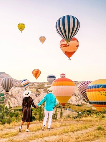 A couple holding hands watches colorful hot air balloons rise over a rocky landscape at sunrise.