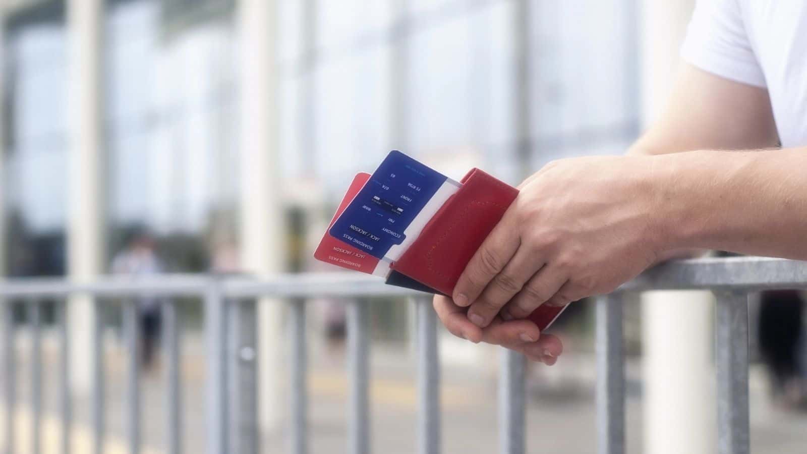 Person holding a red passport holder with a boarding pass inside, standing by an outdoor railing.