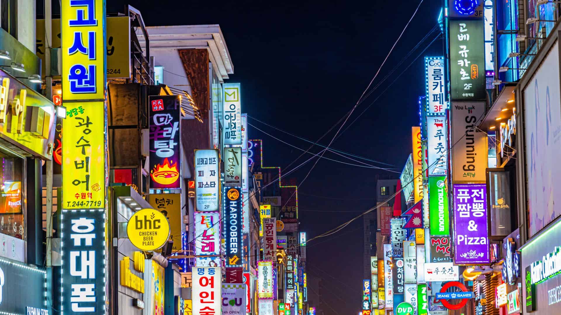 Colorful neon signs with Korean text light up a busy street at night in an urban city scene.