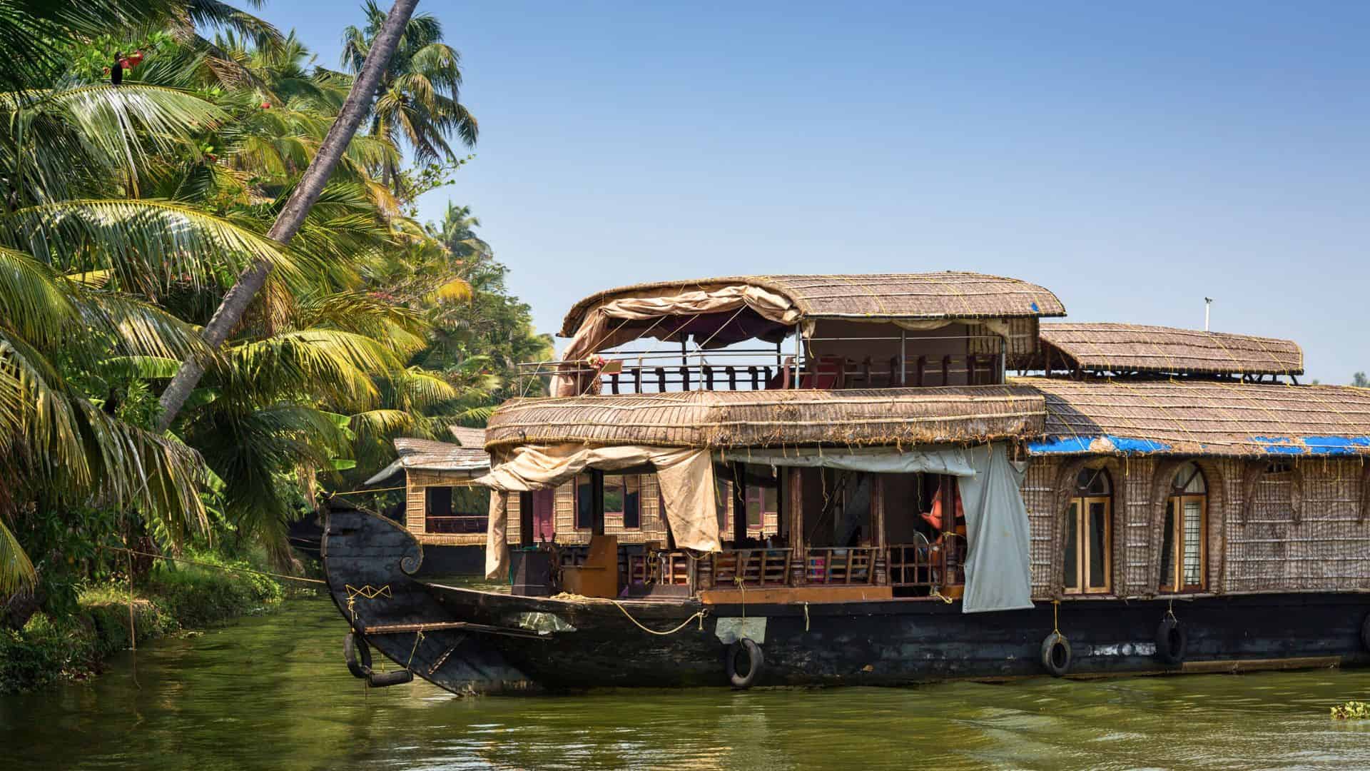 A traditional houseboat floats on a lush, palm-lined river under a clear blue sky.