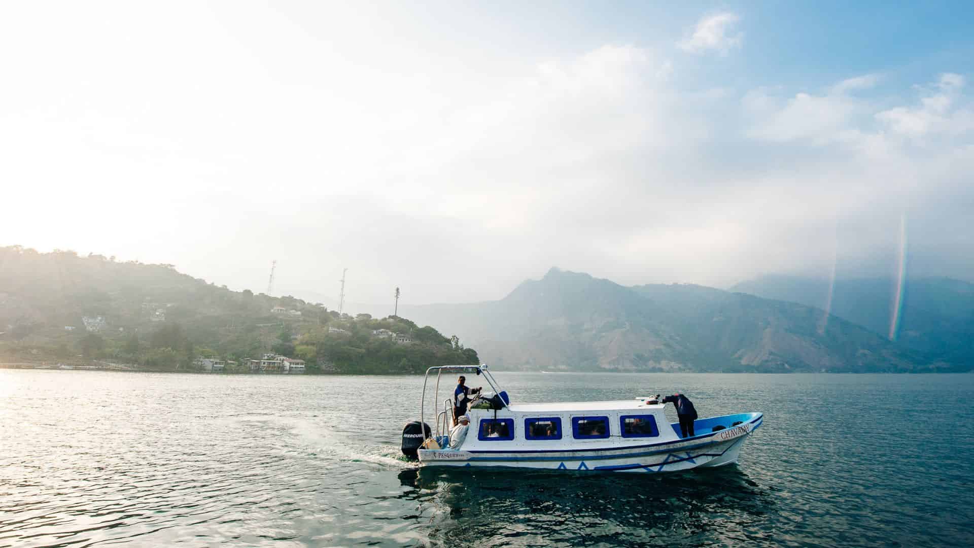 A small white boat cruises on a lake with mountains and a village in the background under a partly cloudy sky.