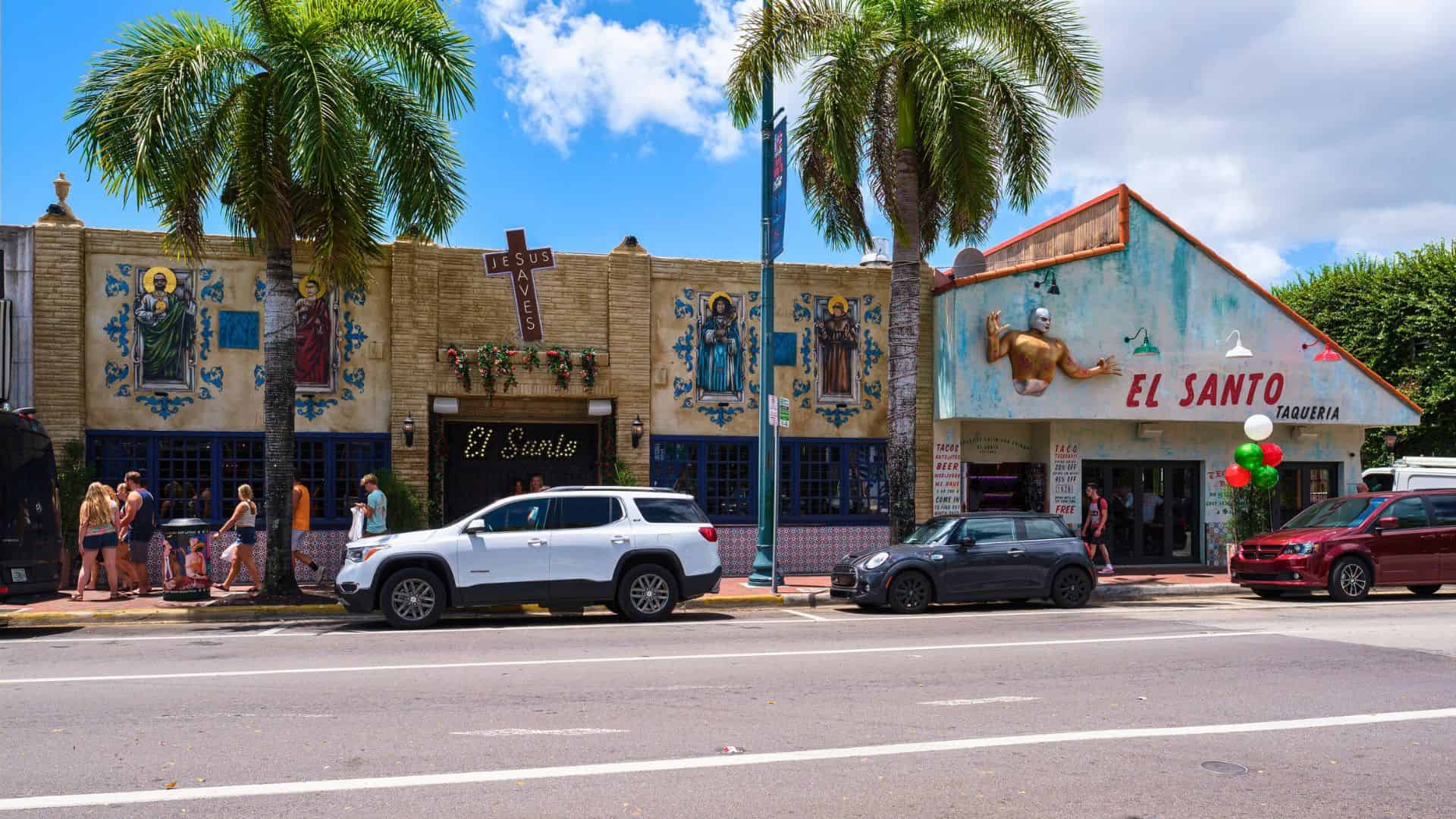 People stand in line outside a colorful taqueria and bar with palm trees and cars in front.