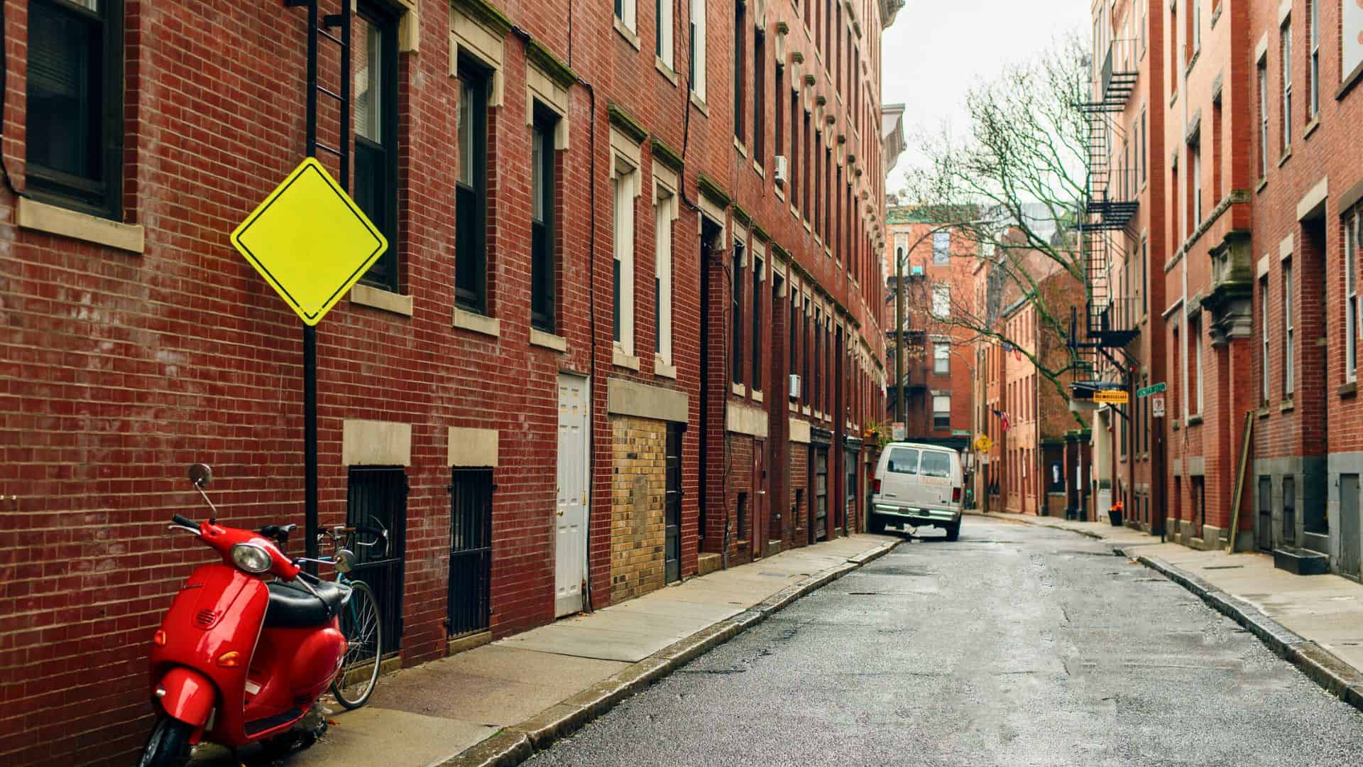 A red scooter and a yellow diamond sign beside a wet, empty street lined with red brick buildings.
