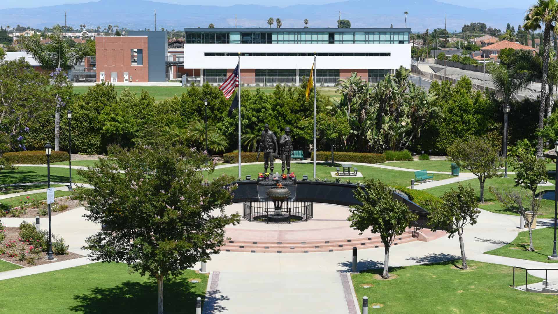 Sunny park with a central statue, American flag, green lawn, and a modern building in the background.