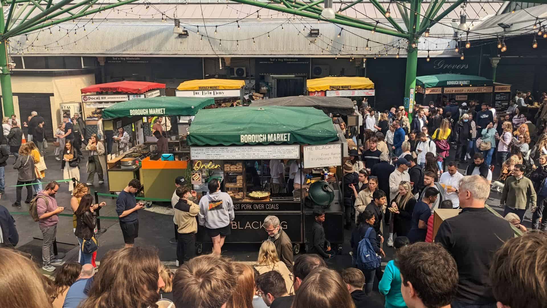 Crowd gathers at Borough Market, with food stalls and people enjoying the outdoor atmosphere under green beams.
