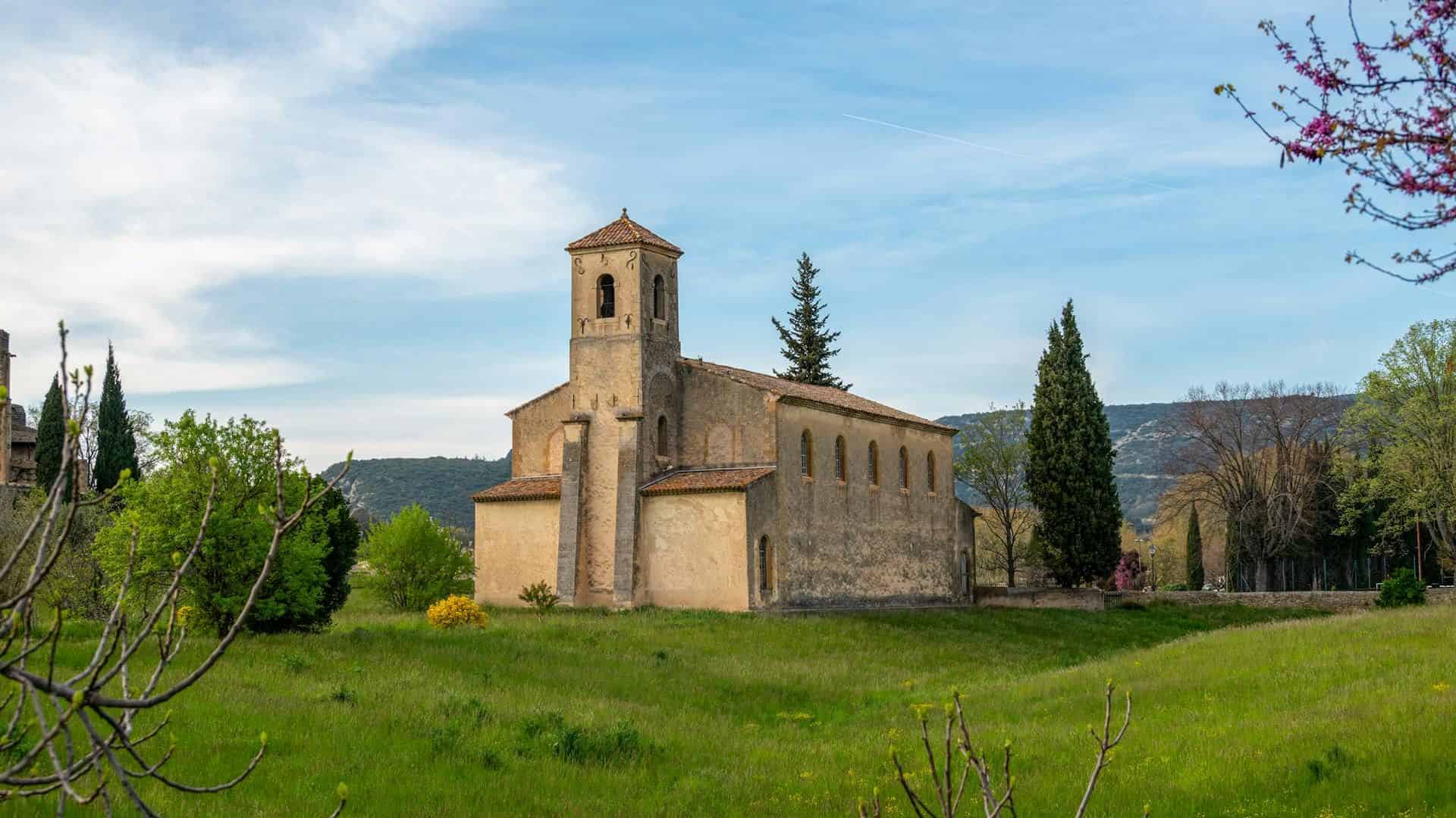 Old stone church with a bell tower surrounded by greenery and trees under a partly cloudy sky.
