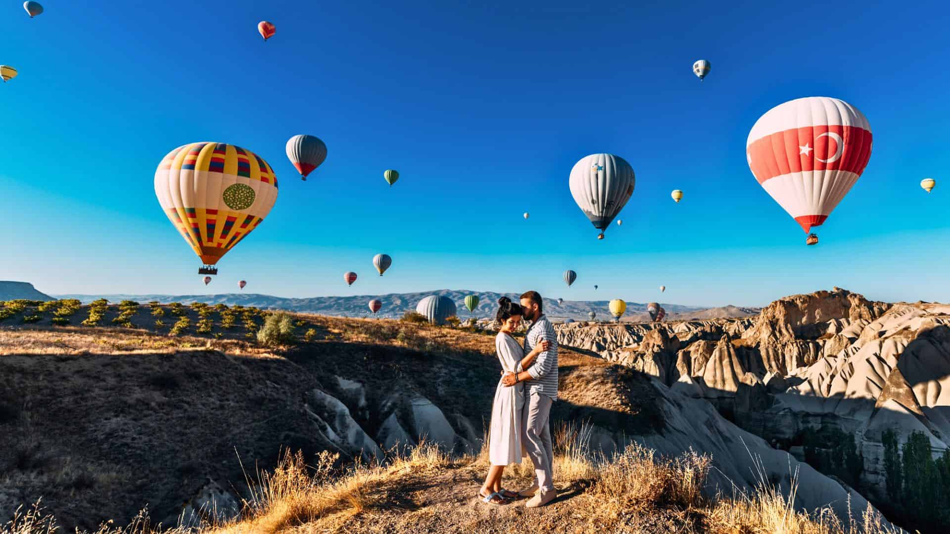 A couple embraces on a cliff as colorful hot air balloons float above a rocky landscape under a clear blue sky.