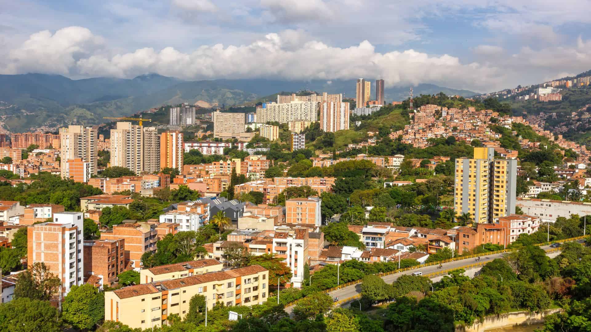 Cityscape with tall buildings, green hills, and mountains under a partly cloudy sky.