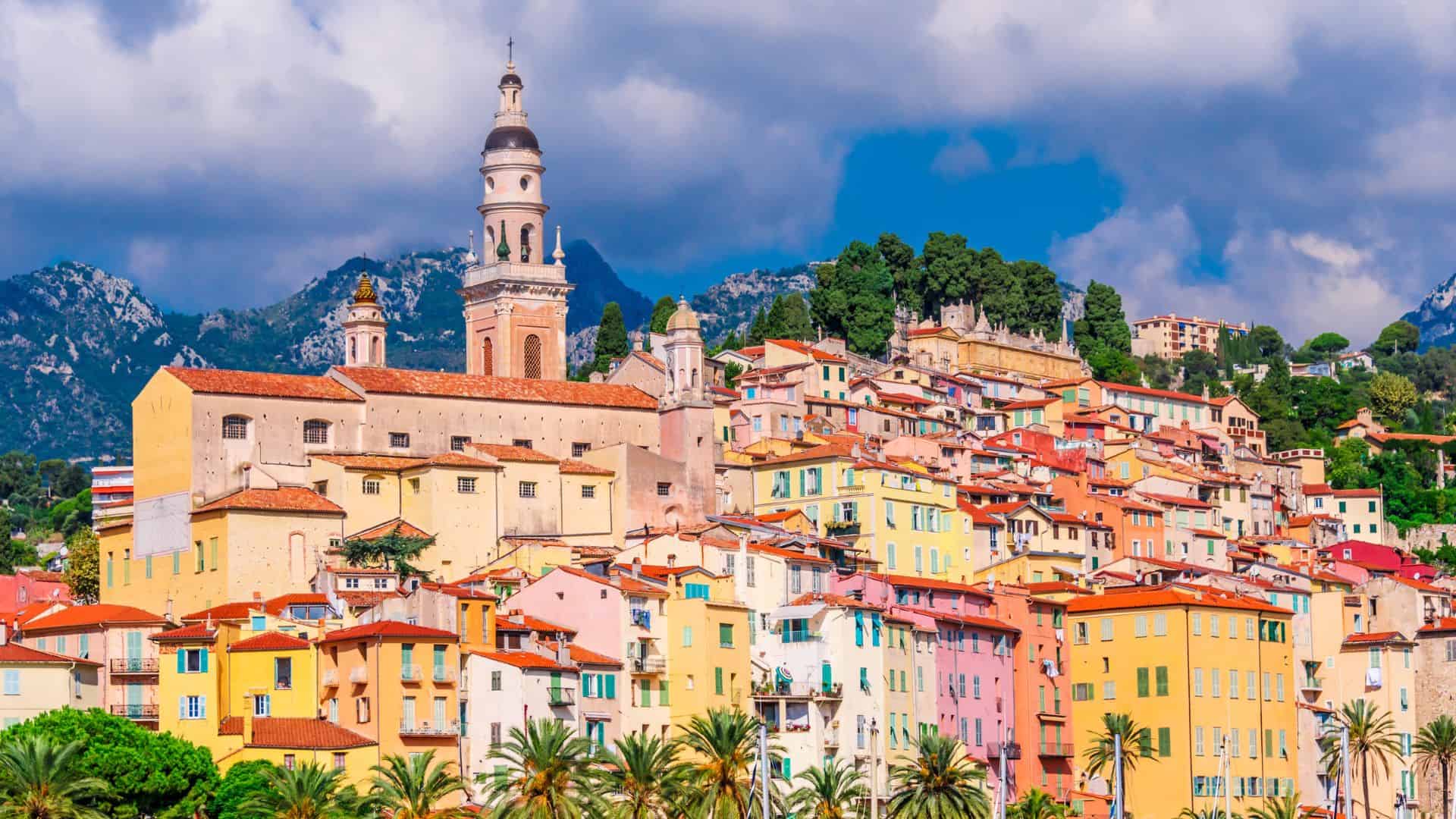 Colorful hillside buildings and a church tower in a Mediterranean town with mountains and palm trees in the background.