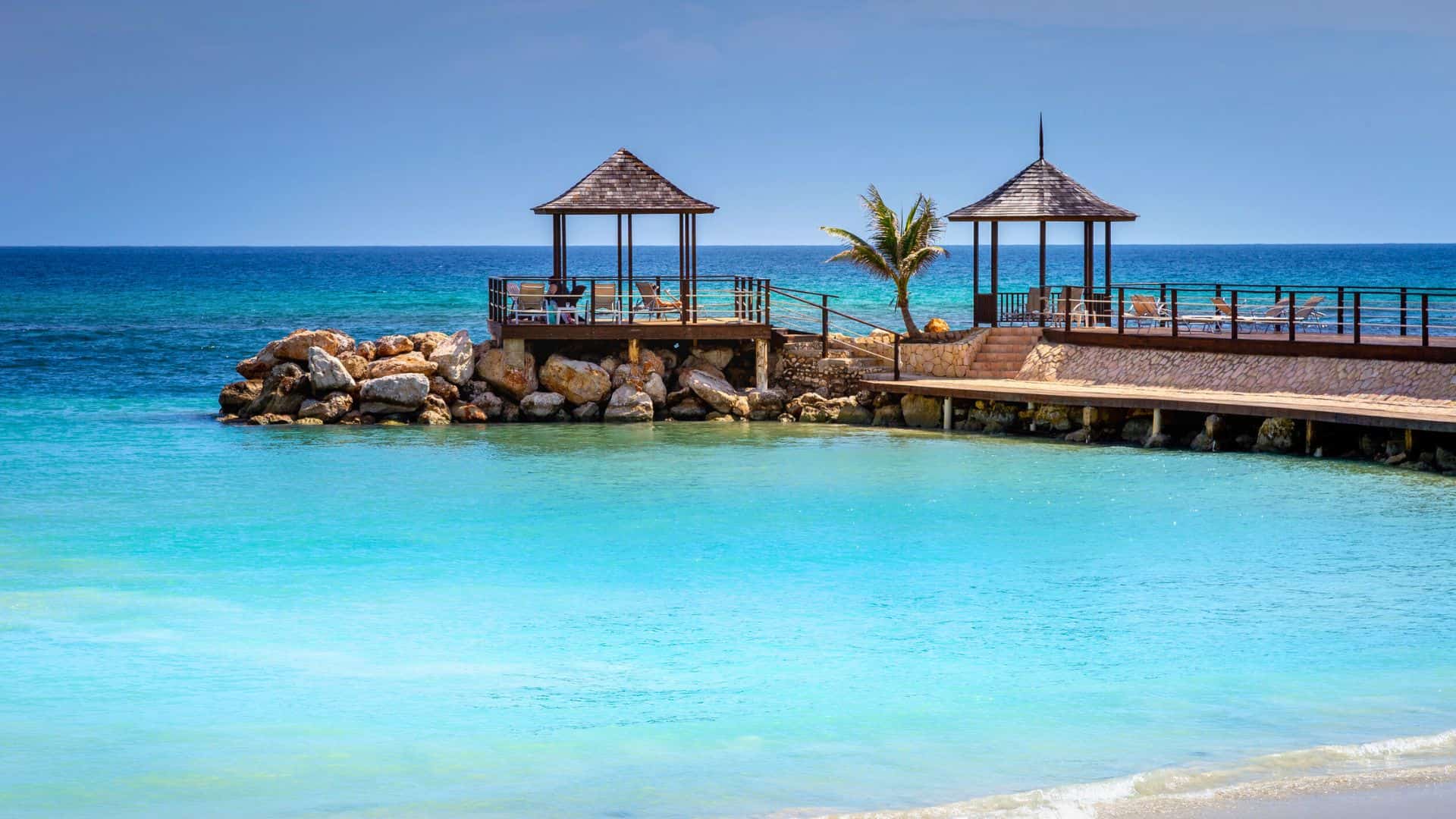 Two wooden gazebos on a rocky pier overlook a calm turquoise sea under a bright blue sky.