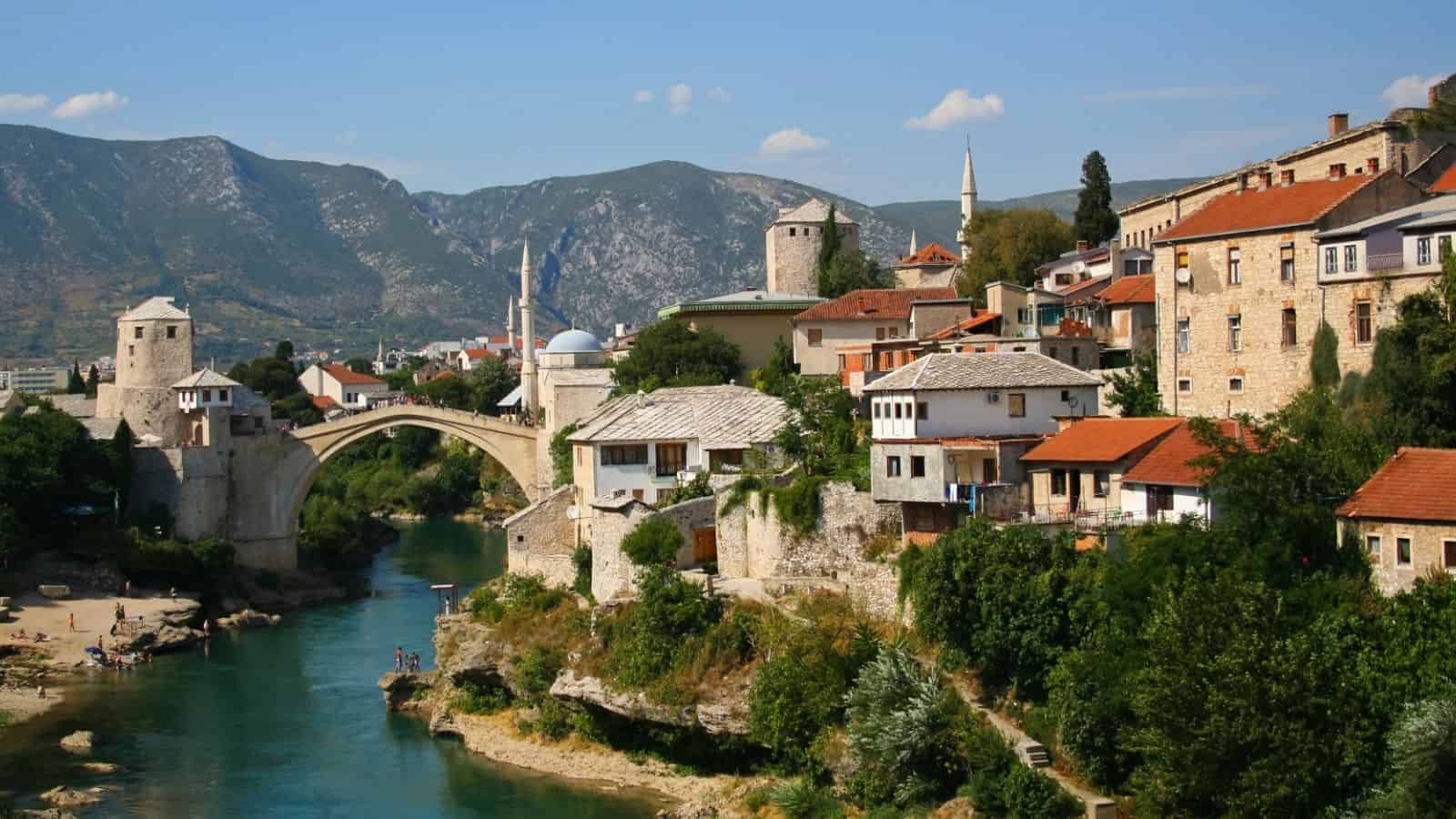 A stone arch bridge spans a river beside historic buildings in a scenic, mountainous town.
