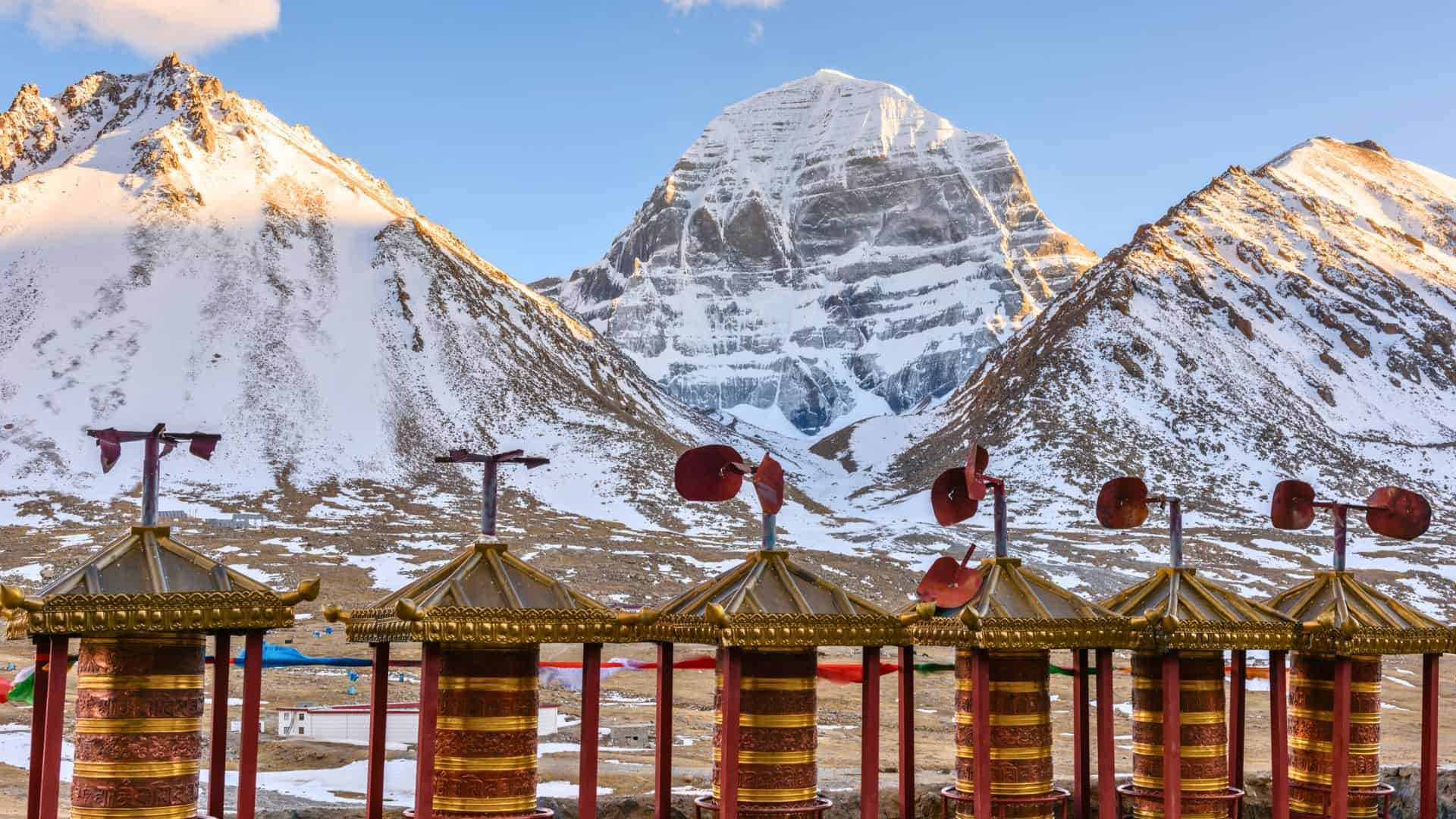 Prayer wheels in front of snow-capped mountains, with Mount Kailash in the background under a clear sky.