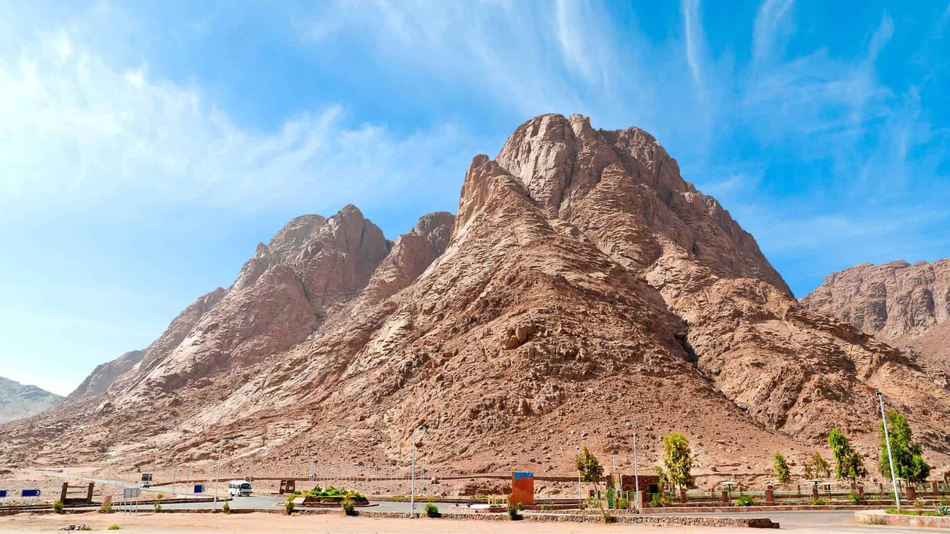 Rocky mountain with a clear blue sky above and a parking area with some greenery in the foreground.