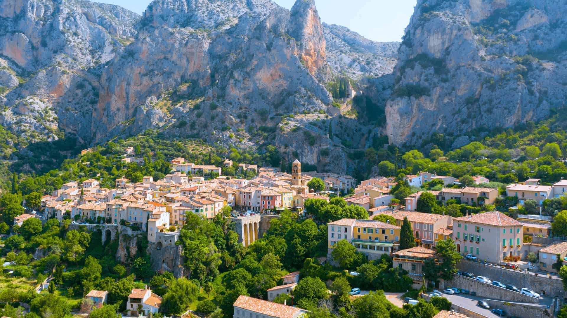 A colorful village nestled among green trees and rocky mountains under a clear blue sky.